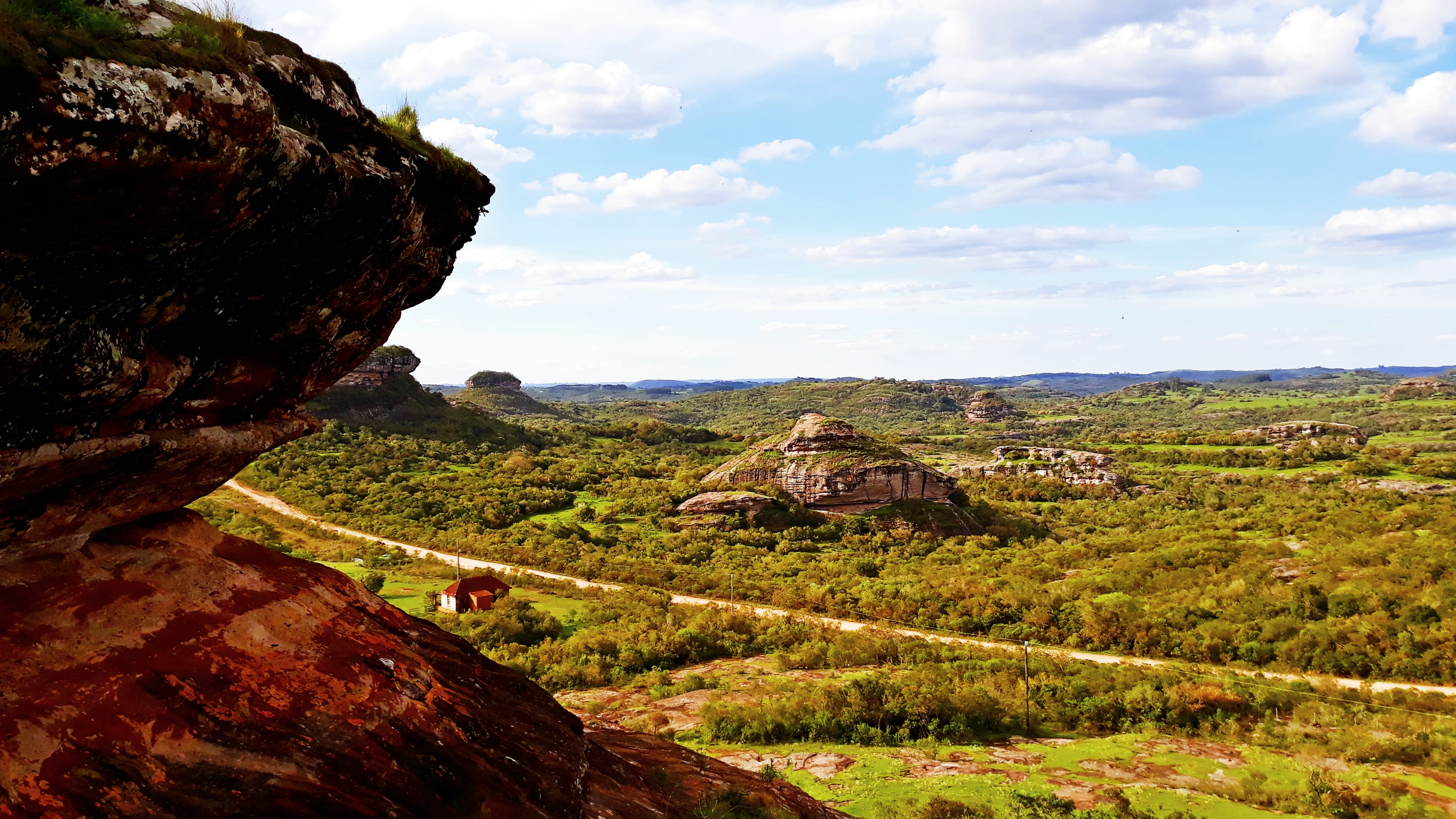 Foto colorida do Geoparque de Caçapava do Sul com rocha à esquerda e paisagem com campos formações rochosas ao fundo