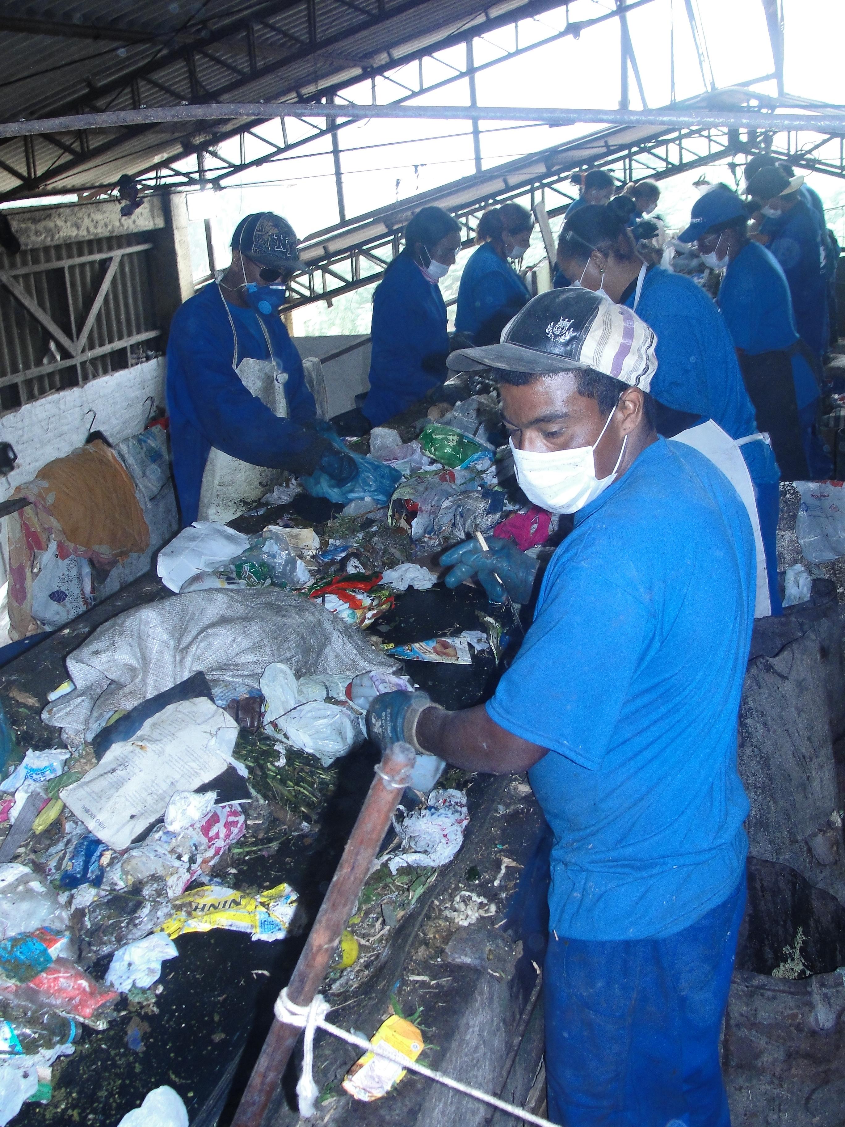 em uma esteira, trabalhadores usam máscaras brancas que tapam nariz e boca e vestem macacões azuis para separar o lixo seco para reciclagem
