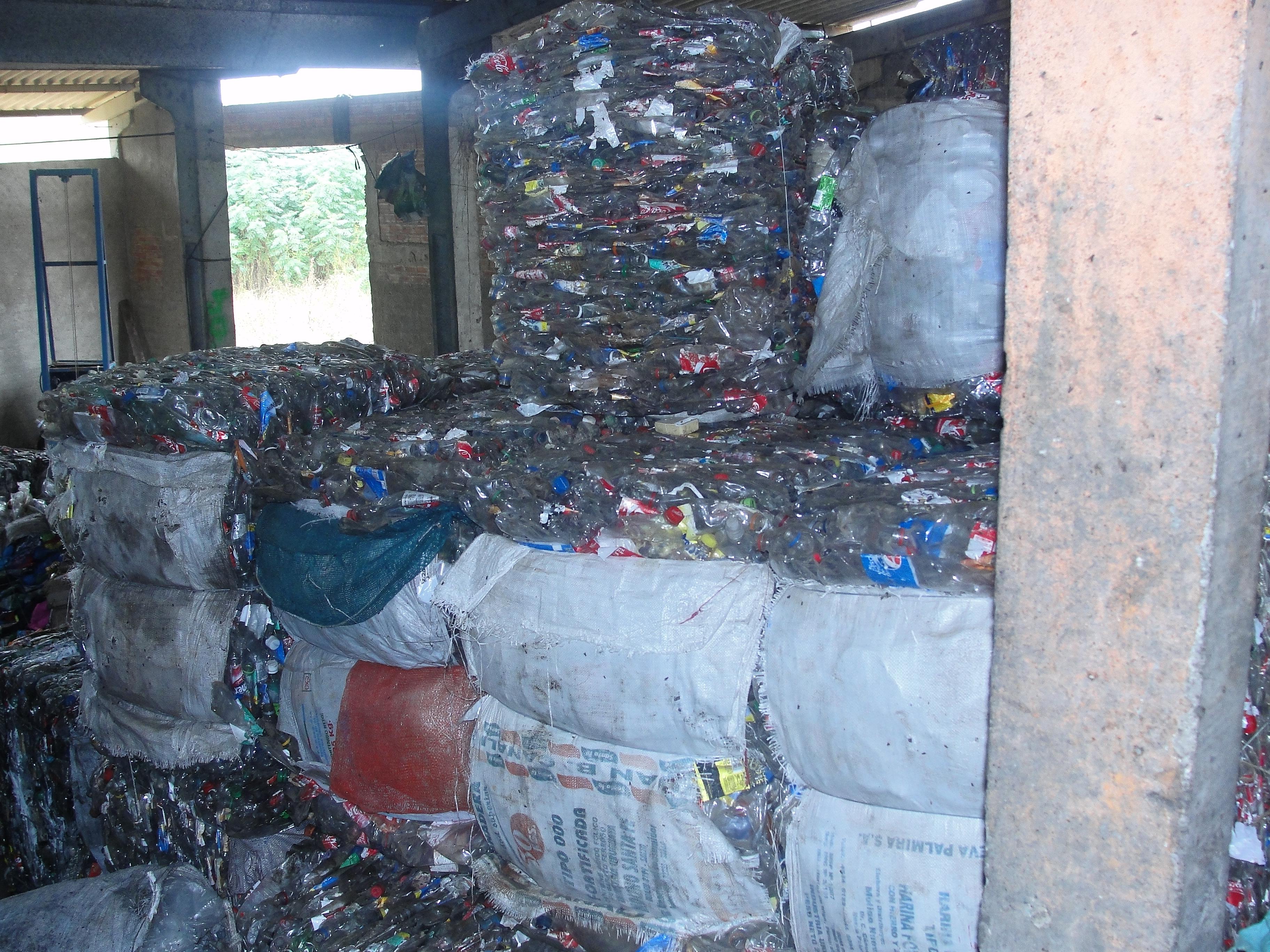 Os materiais compactados para reciclagem estão organizados em fardos grandes, que estão empilhados uns em cima dos outros. Os materiais estão separados por tipos, como latas e garrafas pet