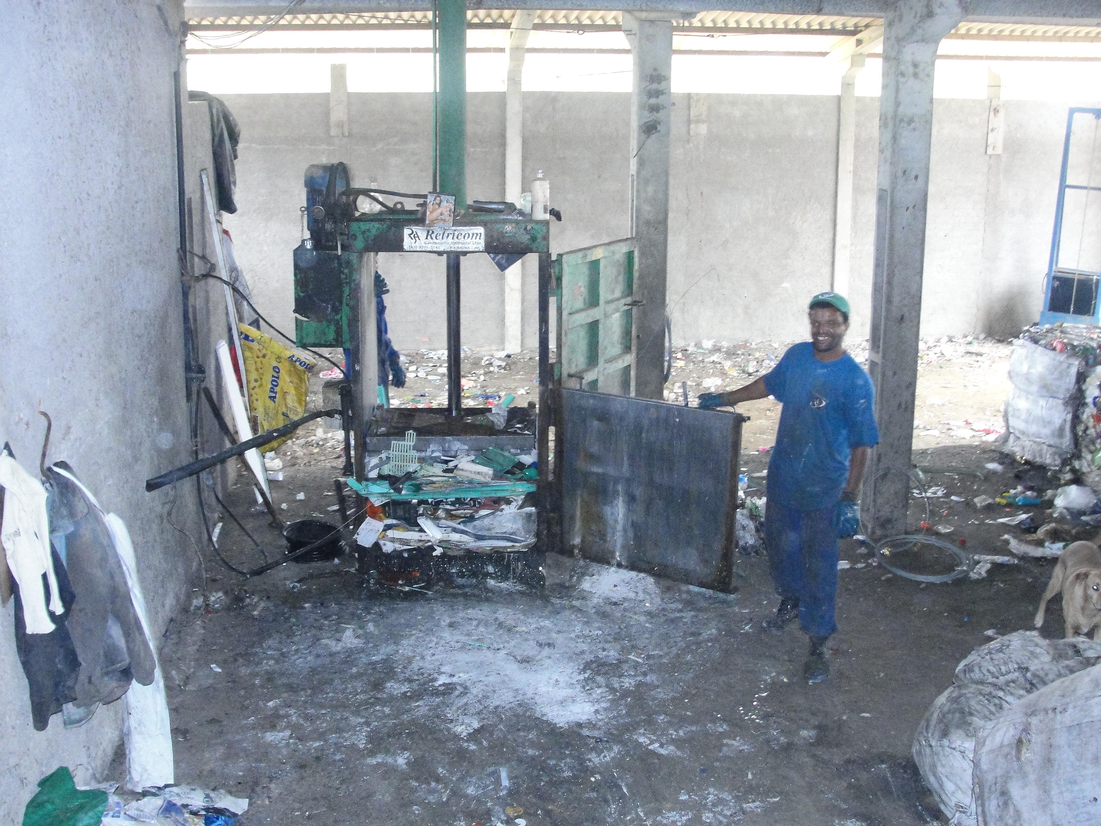 Um homem vestindo macacão azul aciona uma máquina que esmaga e compacta os resíduos secos, facilitando o acondicionamento do material que depois é reciclado