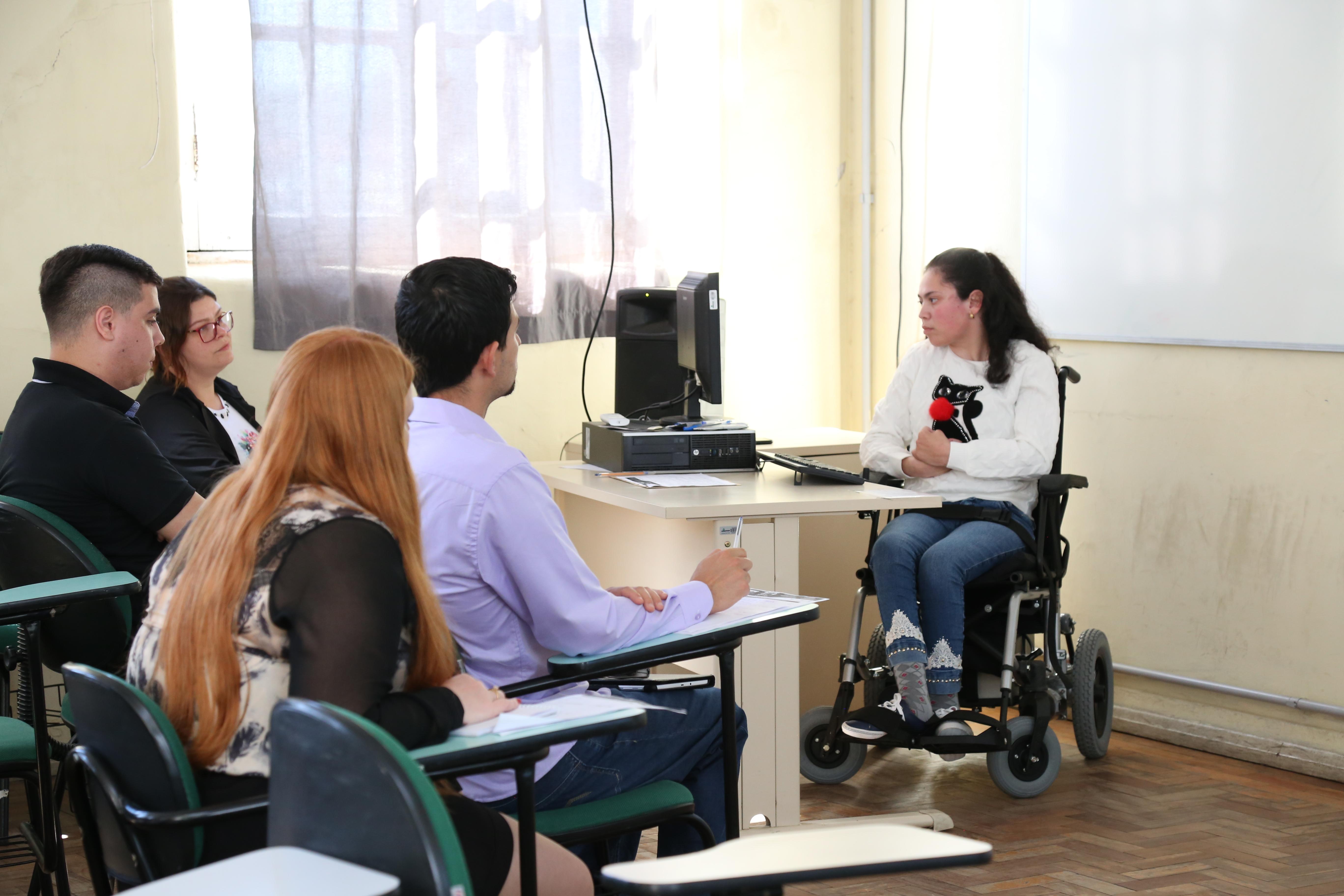 Ao fundo, aluna usuária de cadeira de rodas em frente ao computador. À frente, parcela do público acompanhando a apresentação.