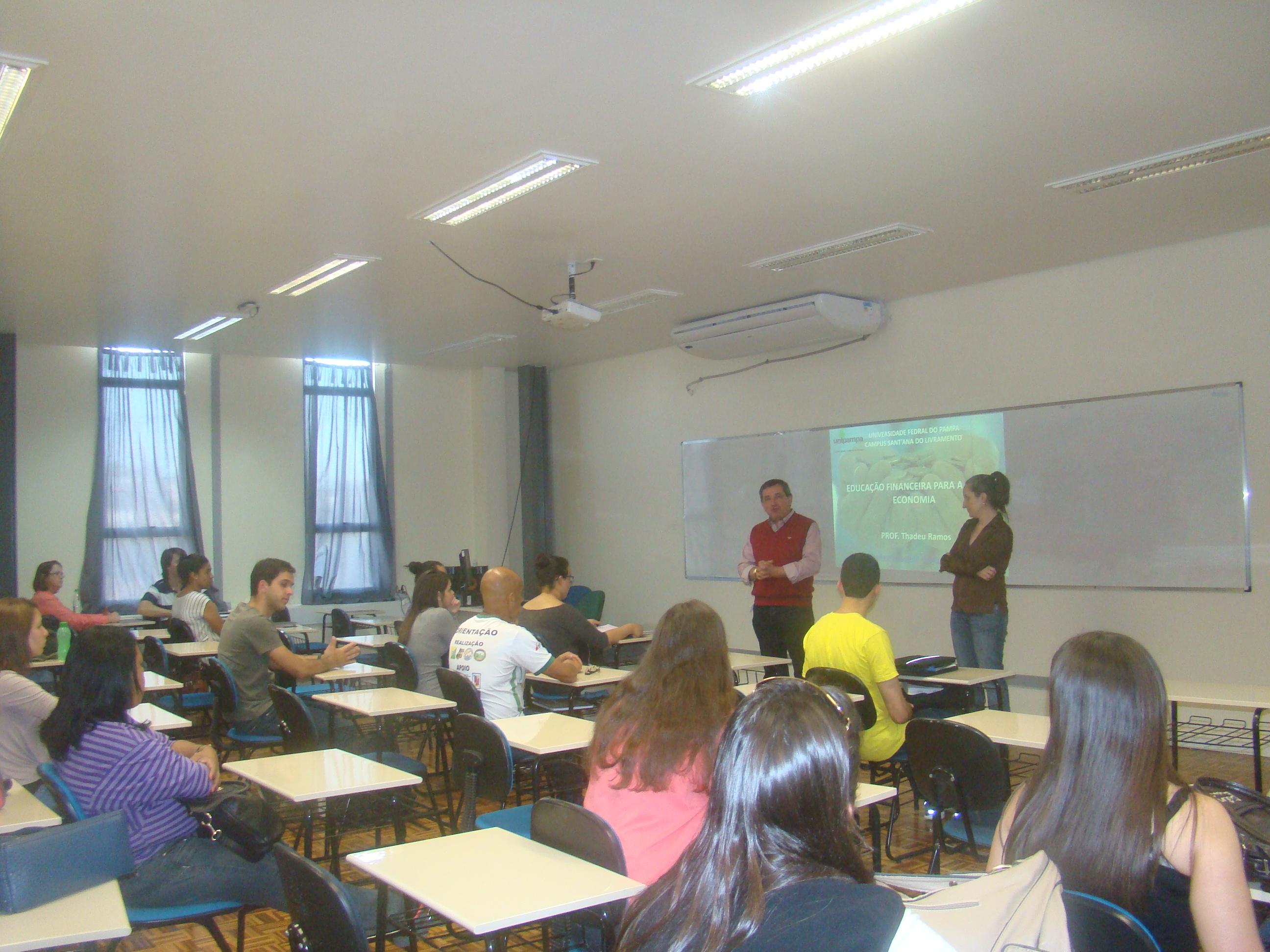 alunos sentados em uma sala de aula olham para dois professores que explicam a proposta do curso, com uma projeção ao fundo.