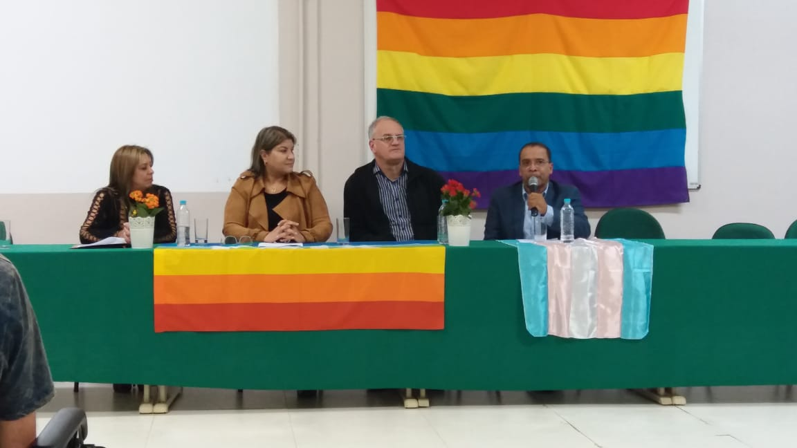 Mesa de abertura do evento com quatro pessoas sentadas. À mesa e ao fundo vê-se a bandeira do arco-íris LGBTQ+