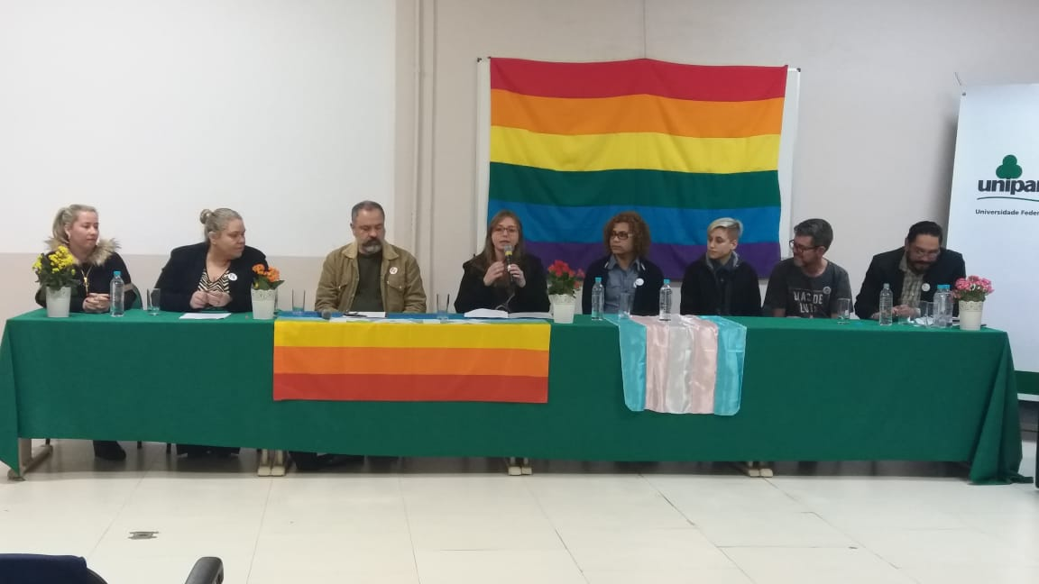 Mesa com debatedores do evento com oito pessoas sentadas. À mesa e ao fundo vê-se a bandeira do arco-íris LGBTQ+