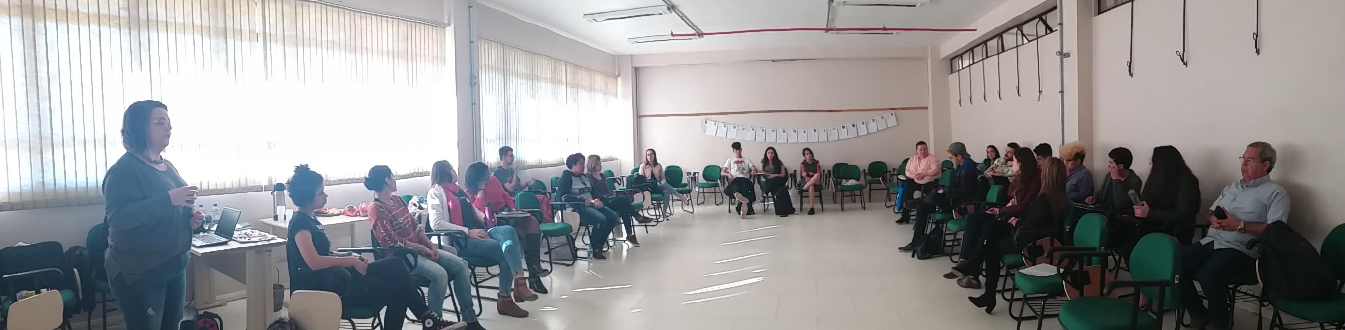 Foto panorâmica de sala com pessoas sentadas organizadas em semicírculo para discussão em grupo