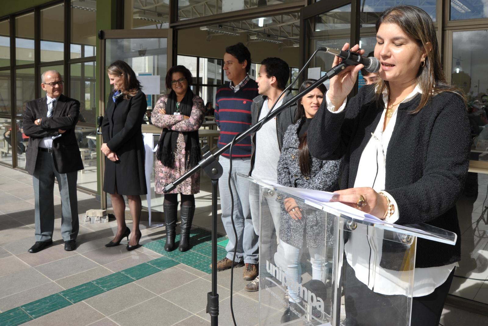 A diretora do Campus Dom Pedrito, professora Nádia Bucco, está em pé, à direita, olha para as pessoas a sua frente e fala ao microfone instalado em um púlpito