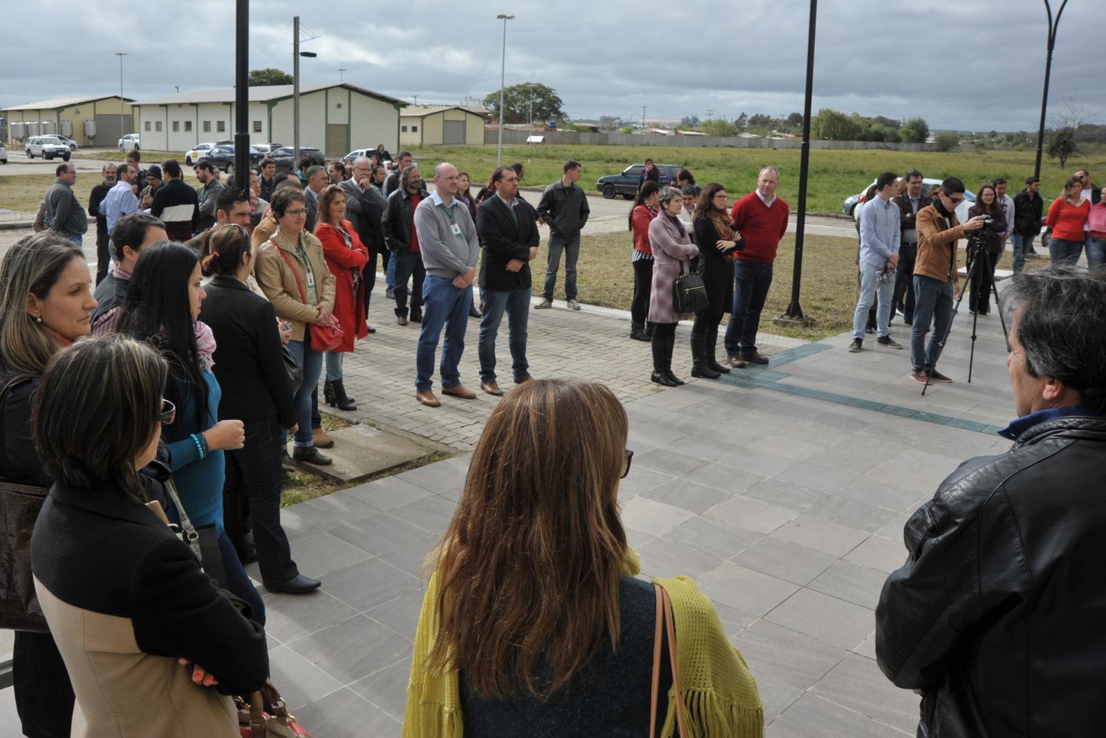 Público em pé, ao ar livre em um dia frio, em frente ao restaurante universitário, acompanham a solenidade de inauguração do serviço