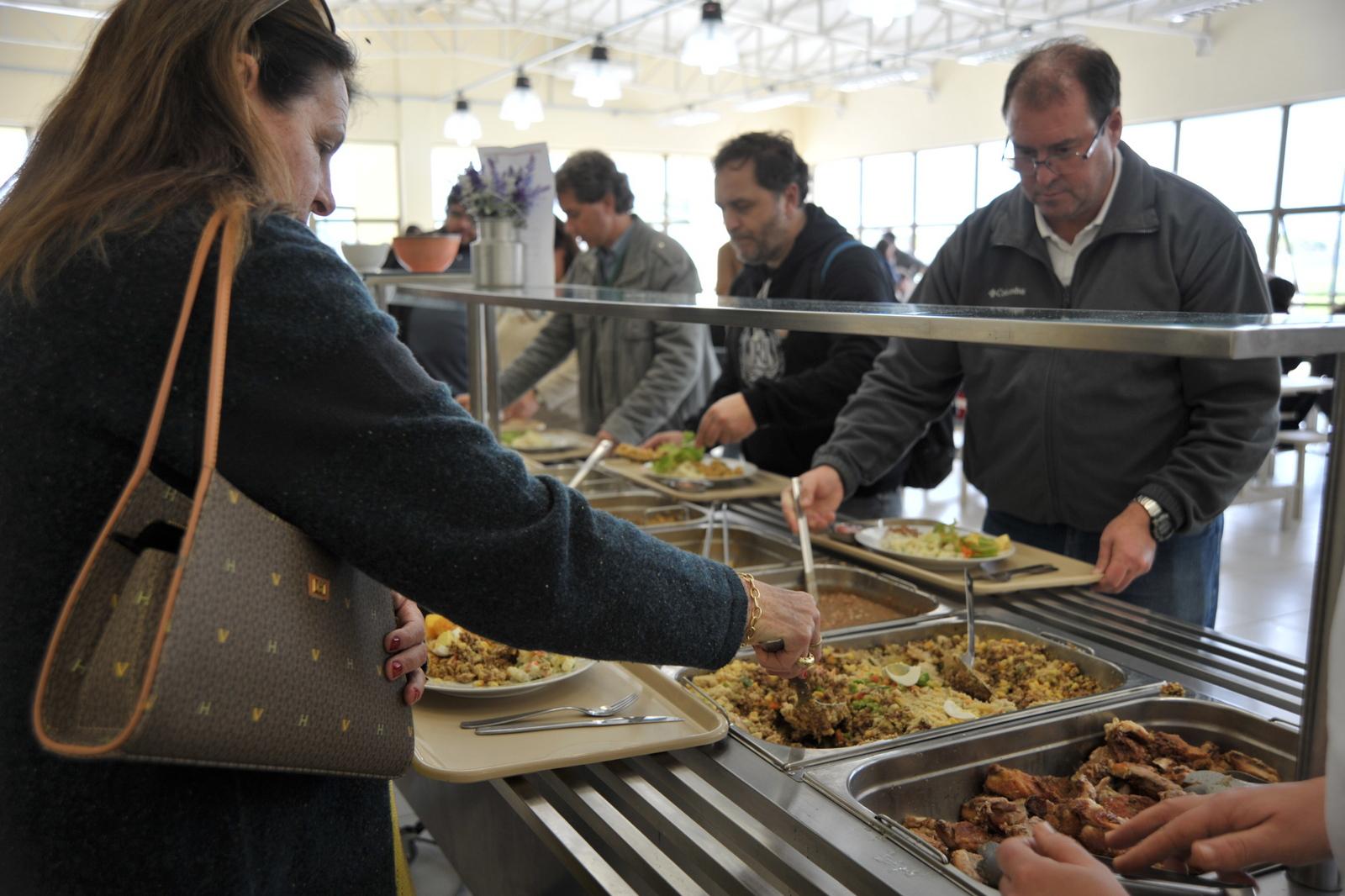 A foto mostra as pessoas servindo seus pratos na linha de servir do restaurante universitário. Uma mulher, com a bolsa a tiracolo, aparece em primeiro plano; no outro lado da linha de servir, mais pessoas servem suas refeições