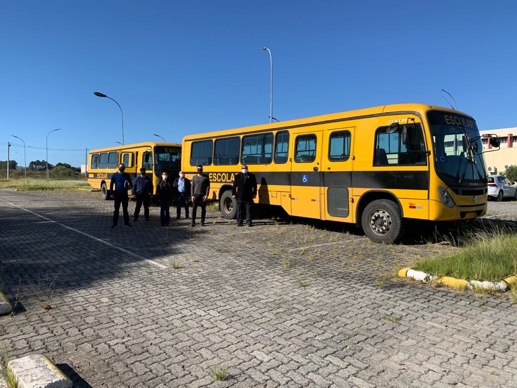 No estacionamento a ceu aberto, pessoas em pé em frente a dois ônibus escolares amarelos