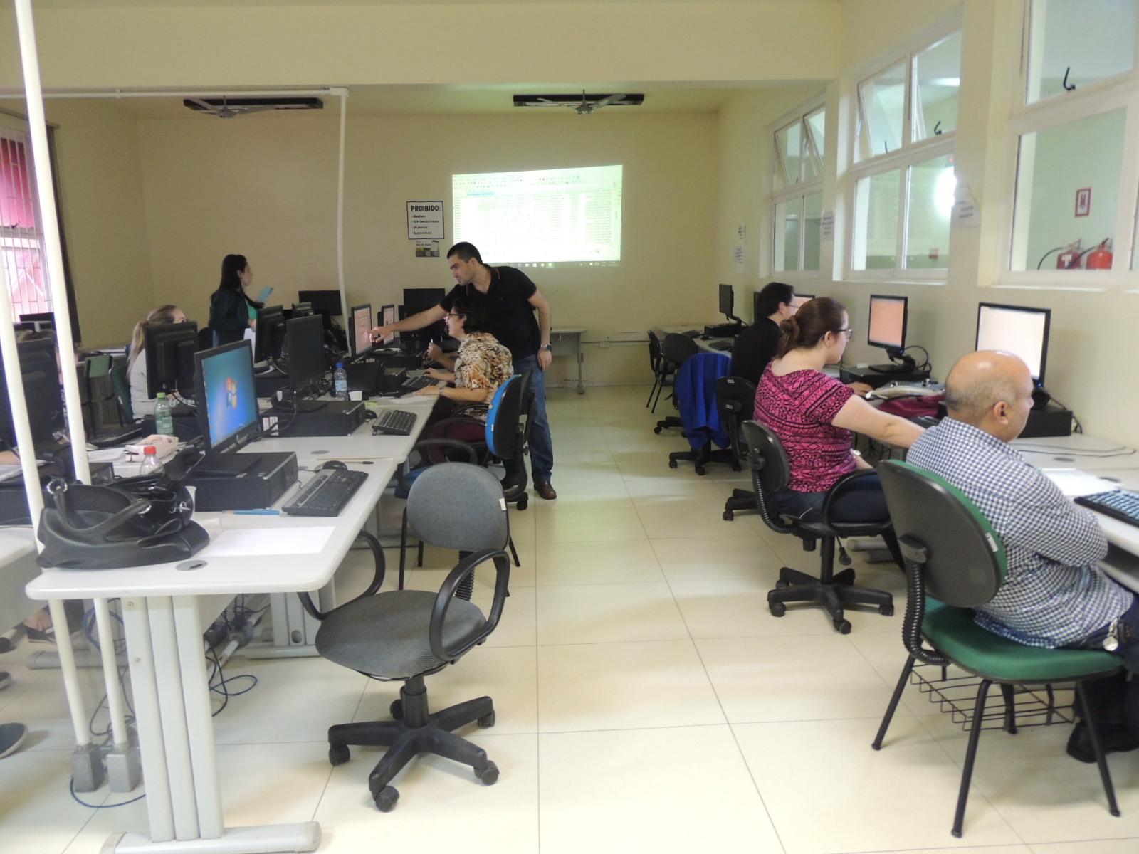 foto mostra uma sala com diversos computadores organizados em estações de trabalho. servidores aparecem sentados trabalhando nessas estações enquanto o instrutor, em pé, passa orientações a uma aluna