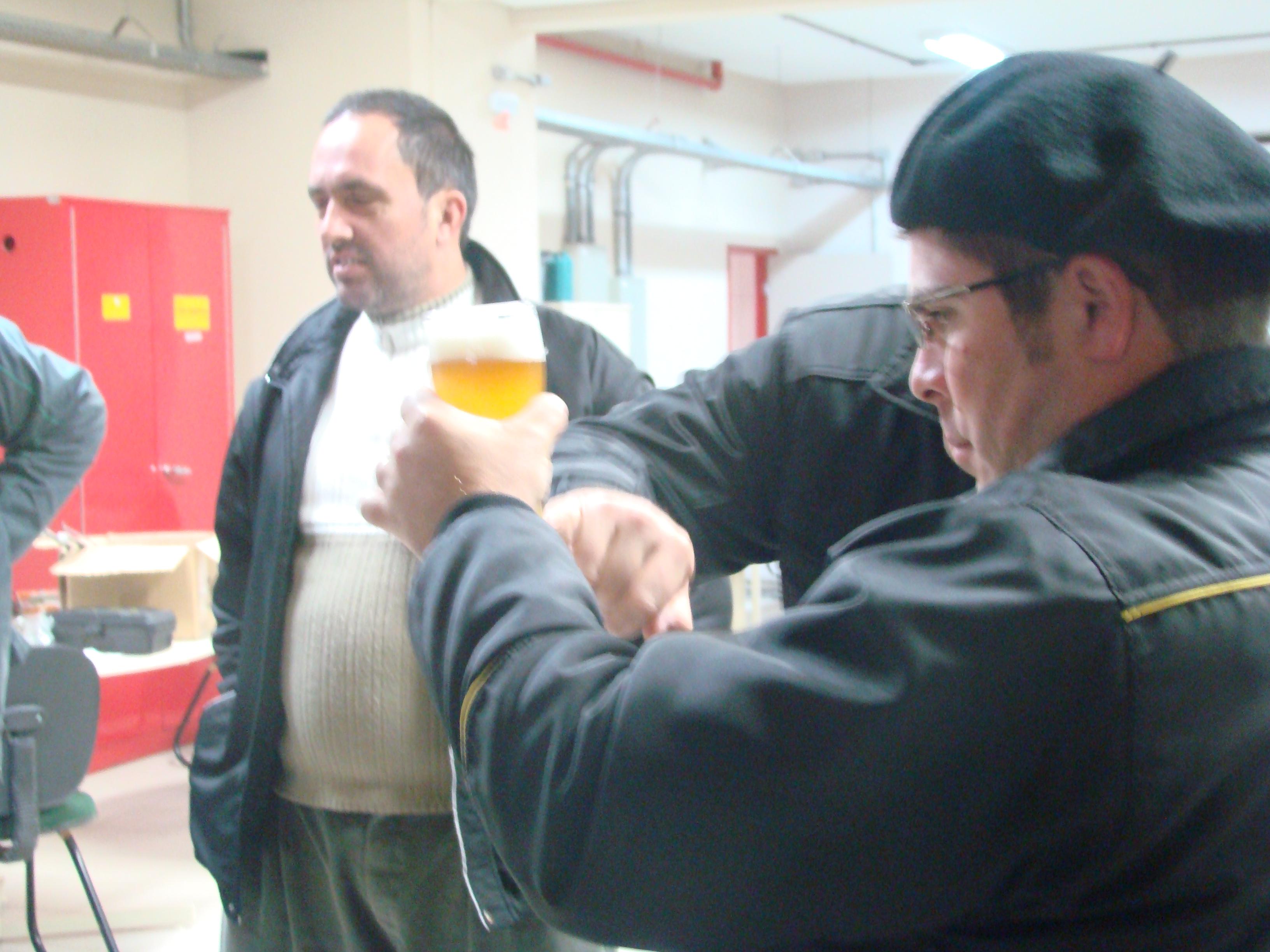 Homem segura copo de cerveja com uma das mãos e a observa. Outro homem figura o fundo da foto.