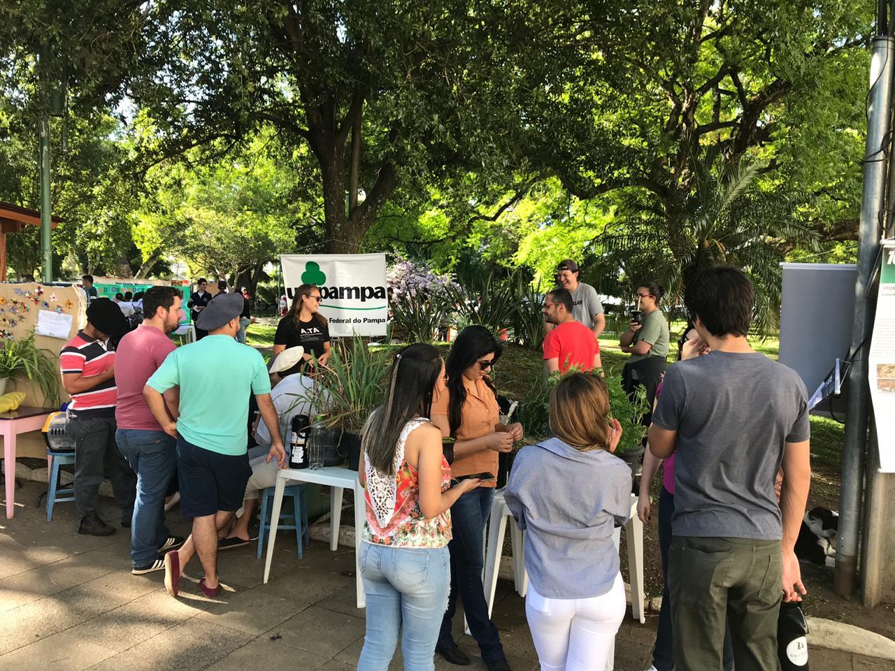 Em uma praça, plantas são expostas em mesas e há um banner com o logo da Unipampa. Cerca de 15 pessoas em pé observam a mostra.