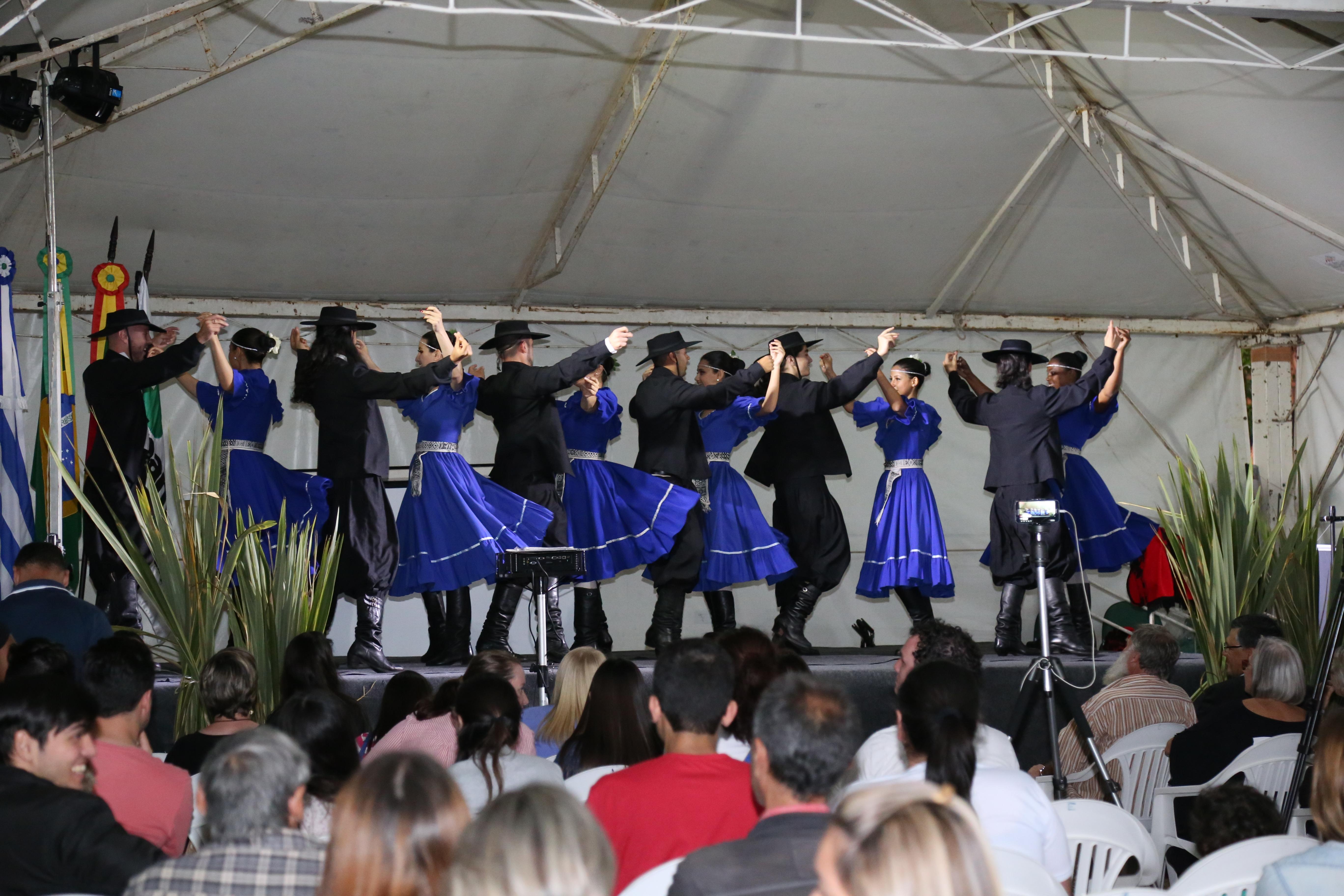 Seis casais dançando no palco usando trajes gaúchos, com cores azul e preto. Público a frente assistindo. 