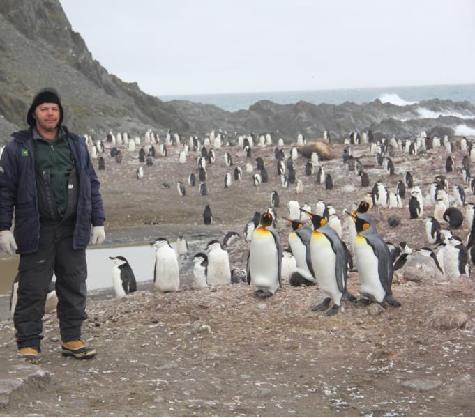 À esquerda da foto um homem em pé, com roupas pretas de frio, casaco grosso, botas para neve, luva, touca. Ao fundo, uma paisagem de montanhas, água e milhares de pinguins. O céu está nublado e o dia está cinza, passando a impressão de baixas temperaturas no local.