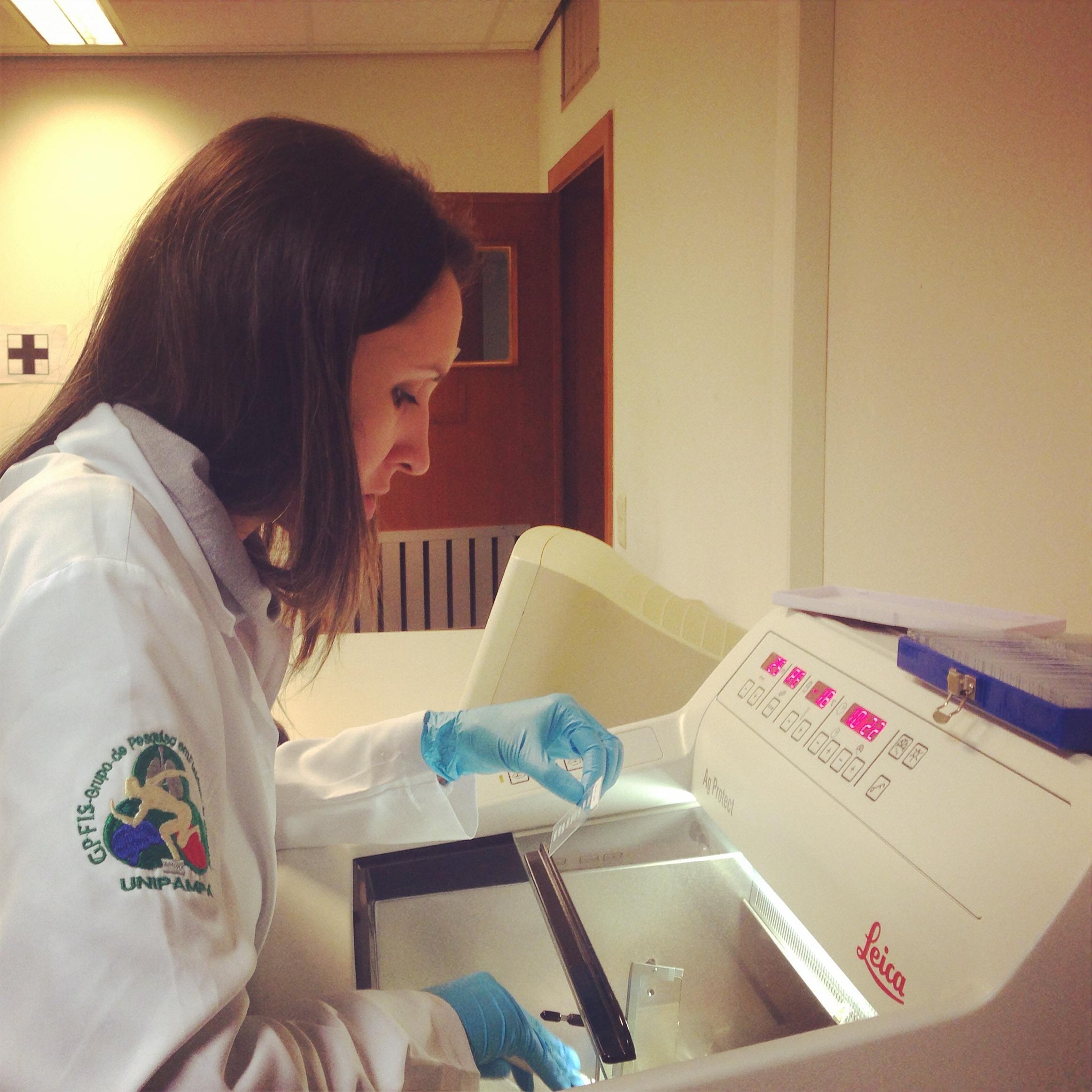 Foto da professora Pâmela Carpes de perfil, sentada de jaleco branco e luvas operando equipamento de laboratório.