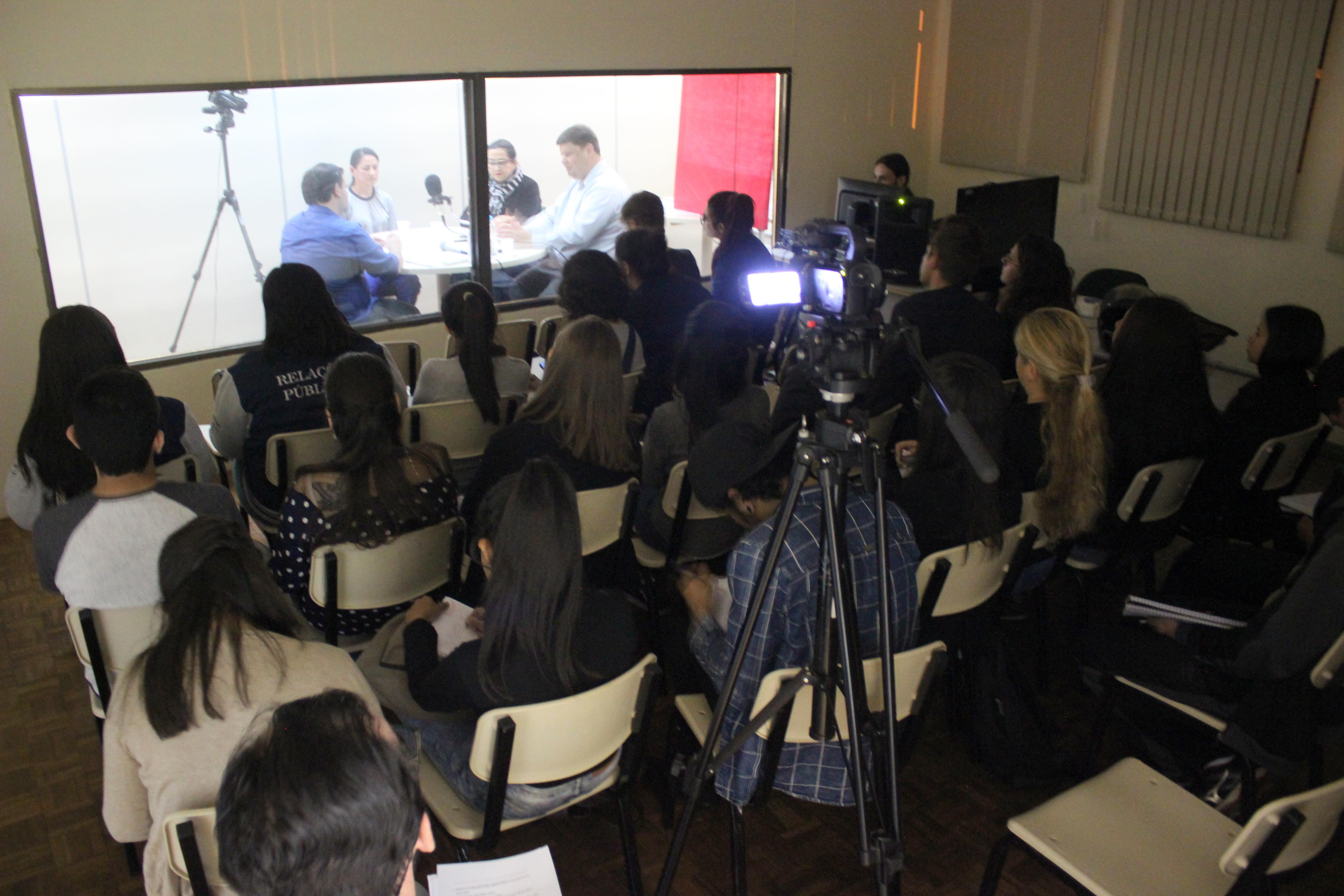 Estudantes em uma sala assistem participantes em grupo focal em outra sala, através de um vidro
