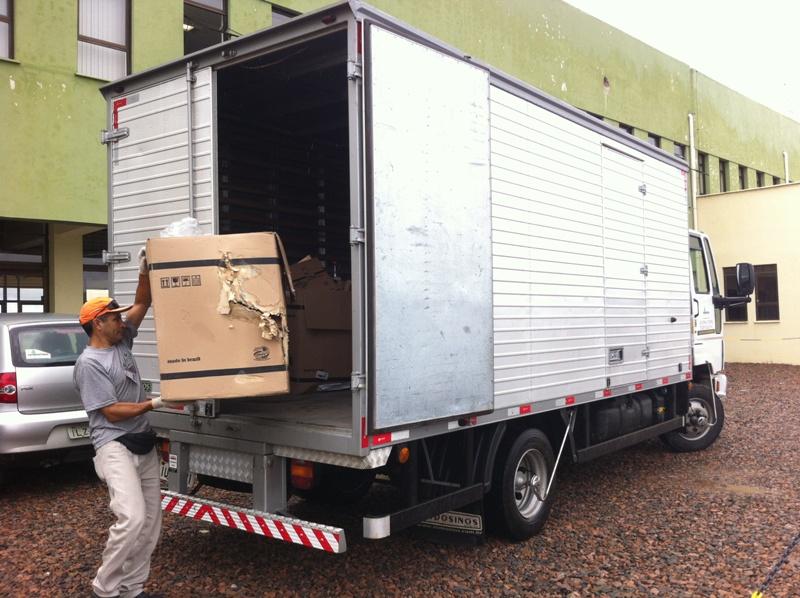 Homem carregando caixas de papelão. Ao fundo um caminhão grande