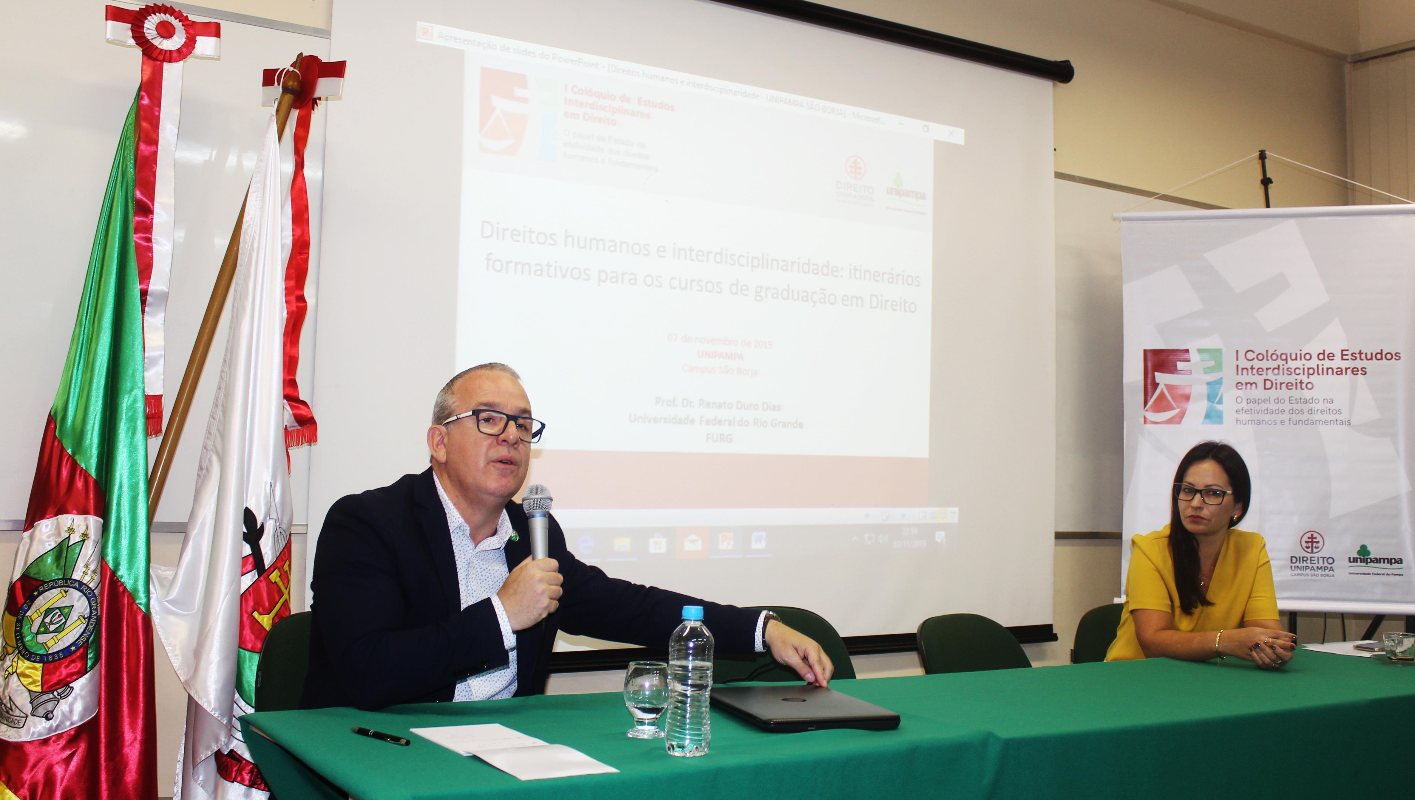 Imagem do professor Renato Duro Dias proferido sua palestra. Na mesa, sentada à sua direita, está a professora Lisianne Ceolin.