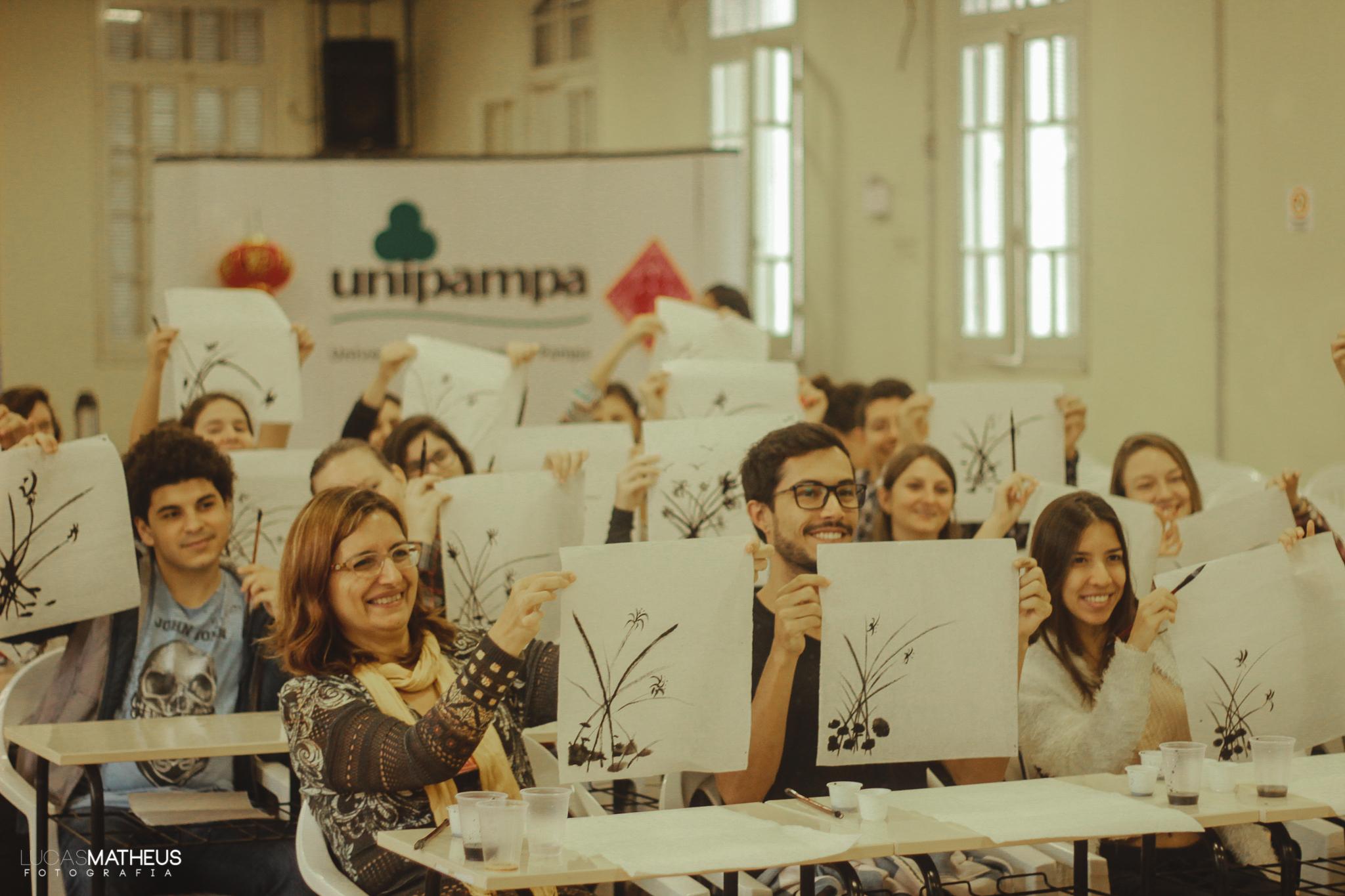 Na imagem um grupo de participantes estão sentados e sorrindo levantam placas com símbolos chineses.